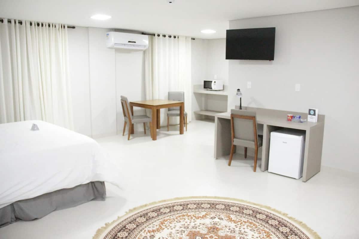 Quarto de hotel com cama, escrivaninha com computador, cadeira, pequeno frigorífico e televisão na parede.