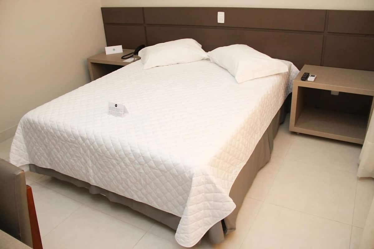 Uma cama bem arrumada com um edredom branco, duas mesas de cabeceira com um telefone e um cartão, e uma cabeceira marrom.