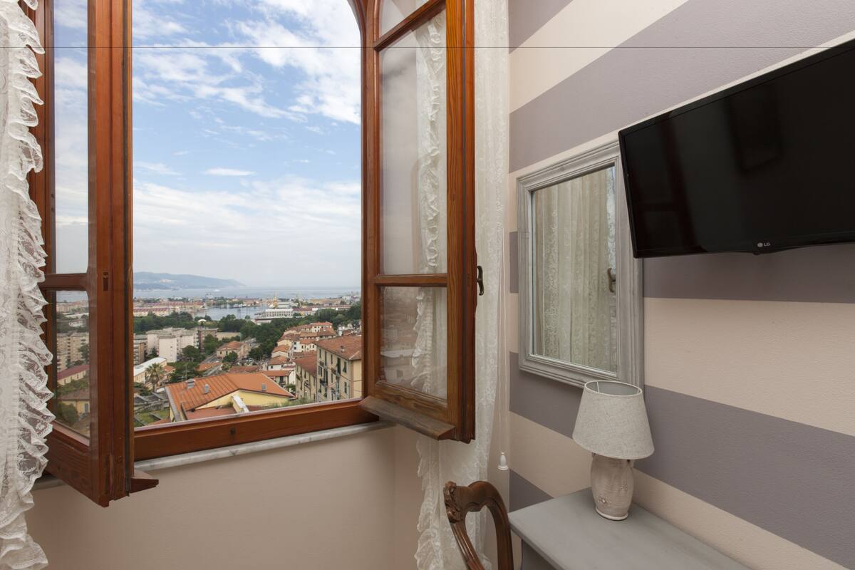 A room with a large window offering a city view, a flat-screen TV mounted on the wall, and a simple bedside table with a lamp.