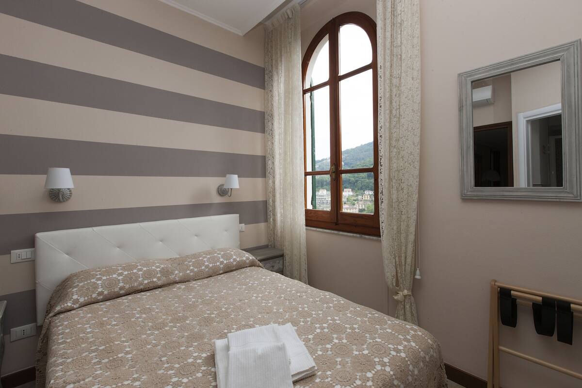 A hotel room with a striped wall, a large window with curtains, a bed with a patterned bedspread, and a bedside table with a lamp.