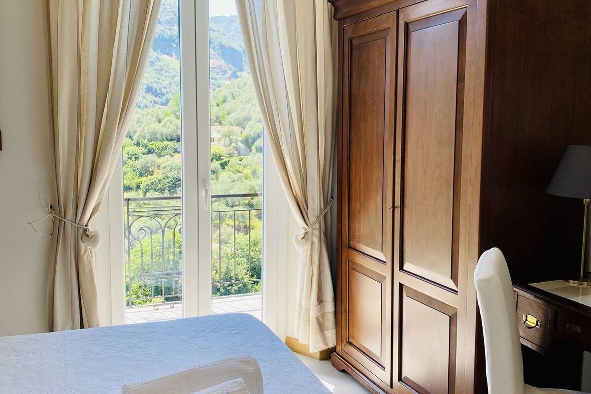 A hotel room with a large bed, a wooden wardrobe, a desk with a lamp, and a view of a balcony with greenery.