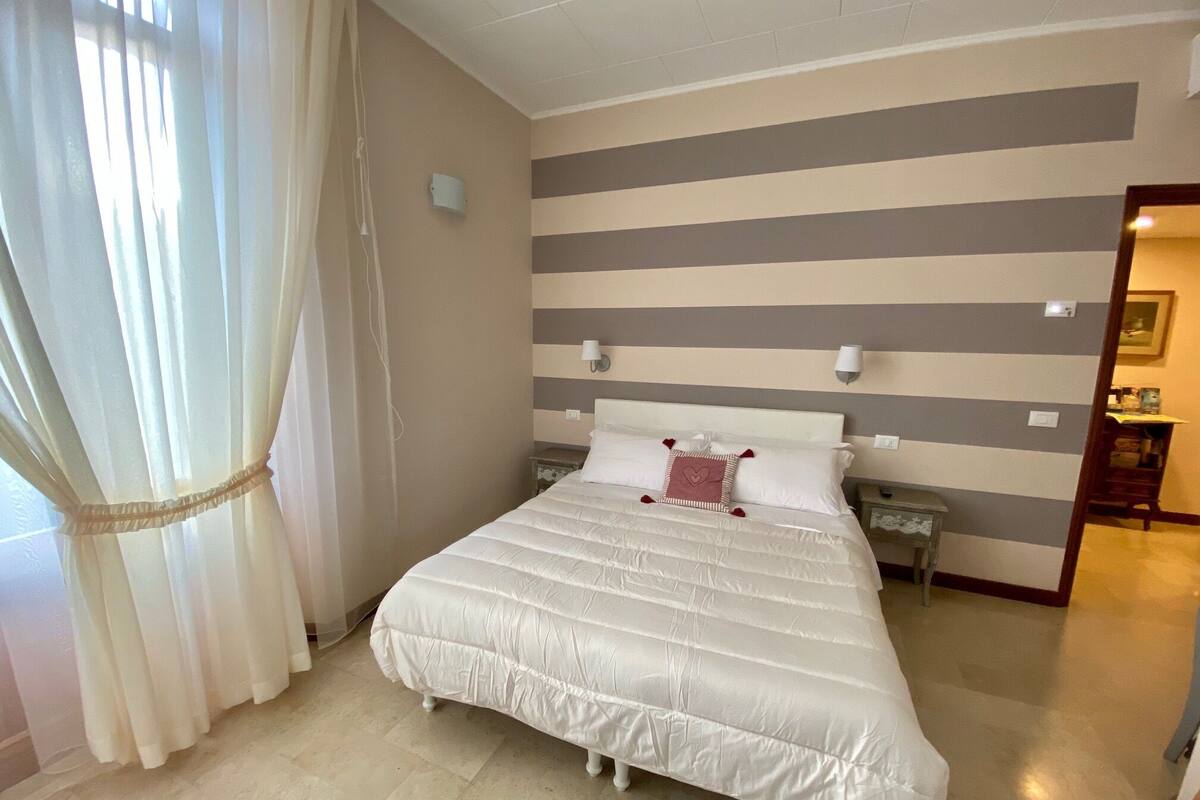 A neatly made bed with a striped wall behind it, a small bedside table, and a curtain on the left.