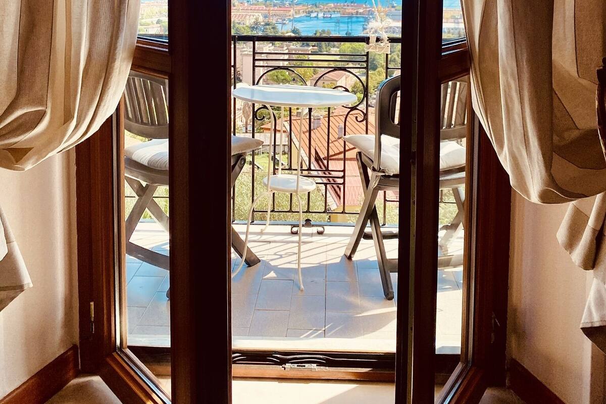 A balcony with a table and chairs, a view of the sea and buildings, and a curtain partially covering the window.