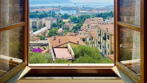 Vista desde una ventana con un arco, que da a un paisaje urbano con edificios, vegetación y una masa de agua distante.