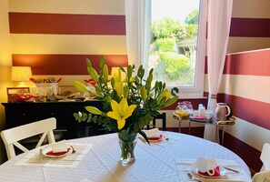 Una mesa de comedor con manteles blancos, un florero con flores amarillas y una ventana con cortinas.