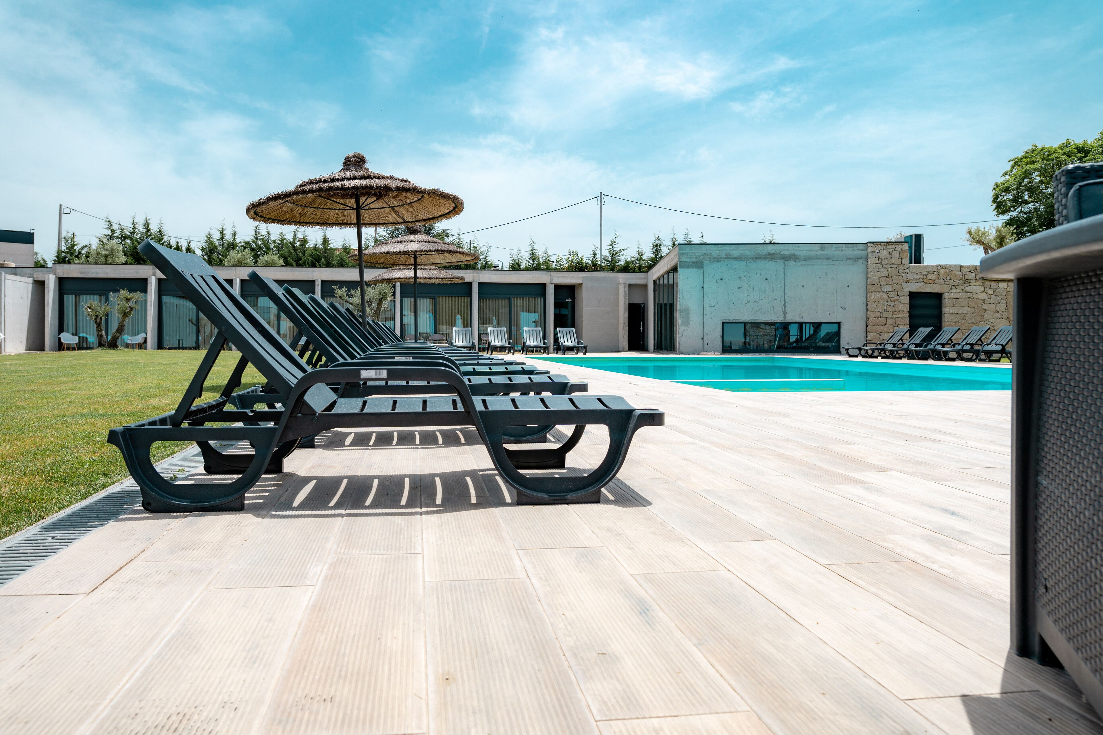 Een zwembadterras met ligbedden en parasols, een houten terras en een modern gebouw op de achtergrond.