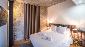 Een moderne hotelkamer met een accentmuur van steen, een groot bed met witte lakens en een nachtkastje met een lamp.