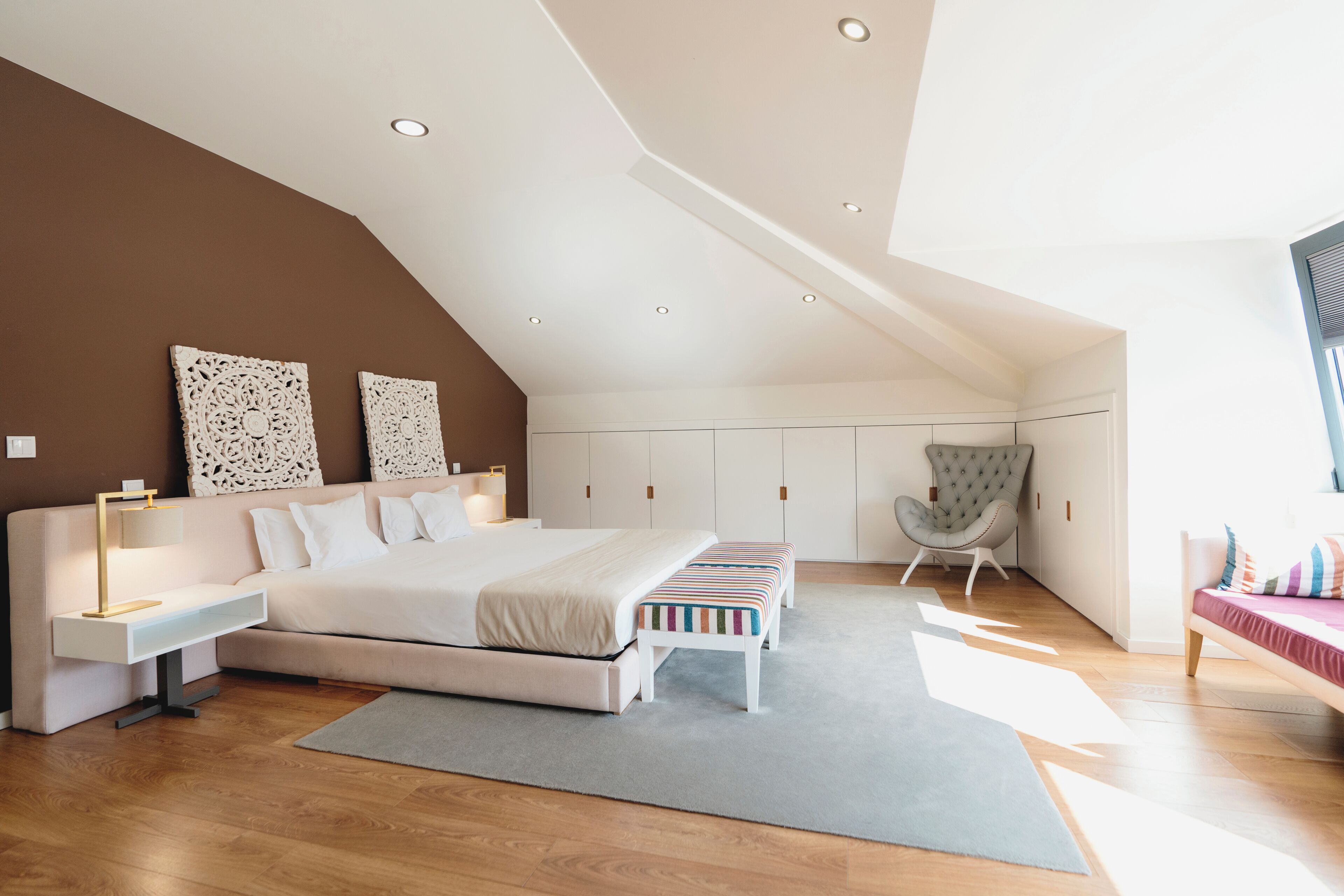 A modern bedroom with a sloped ceiling, a large bed, bedside tables, a chair, and a bench.