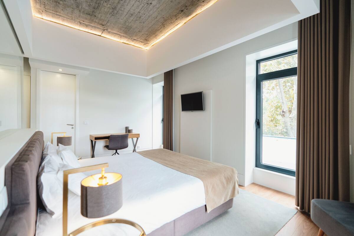 Ein modernes Hotelzimmer mit einem großen Bett, einem Schreibtisch, einem Fernseher und einem Fenster mit Vorhängen.