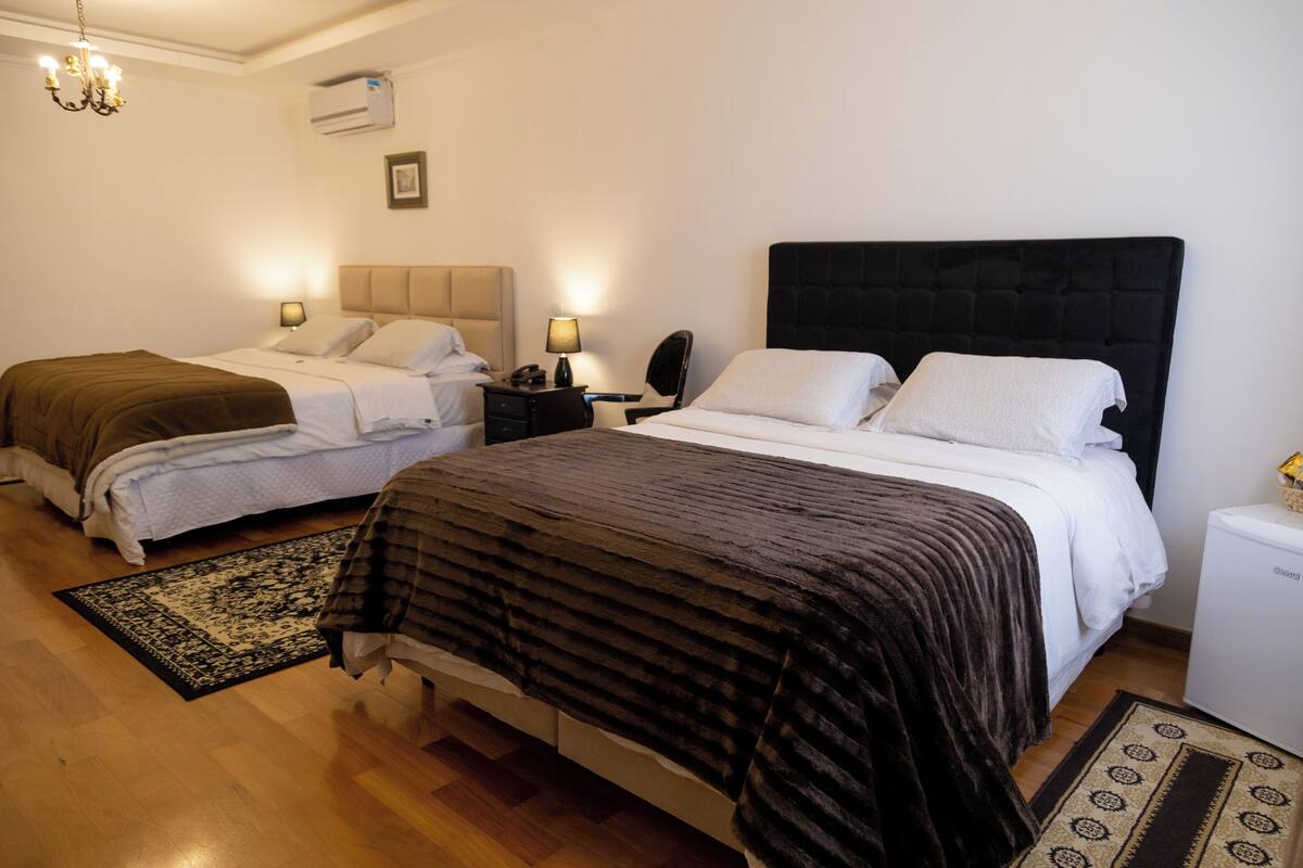 A hotel room with two beds, a small bedside table, a nightstand, a lamp, a heater, and a wall-mounted air conditioner.