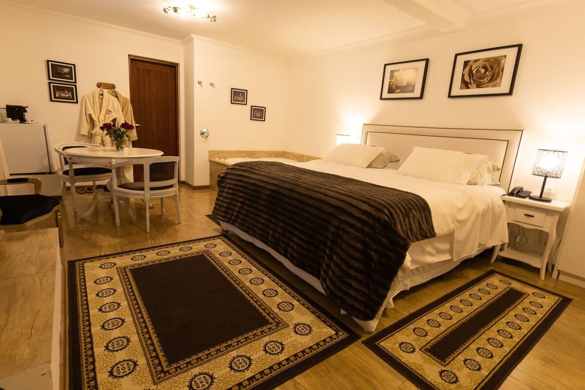 A hotel room with a bed, bedside table, lamp, and a small table with chairs. There are two rugs on the floor, a door, and framed pictures on the wall.