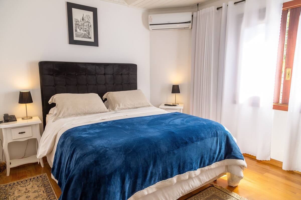 A bedroom with a large bed, white walls, a framed picture, an air conditioner, and a window with curtains.
