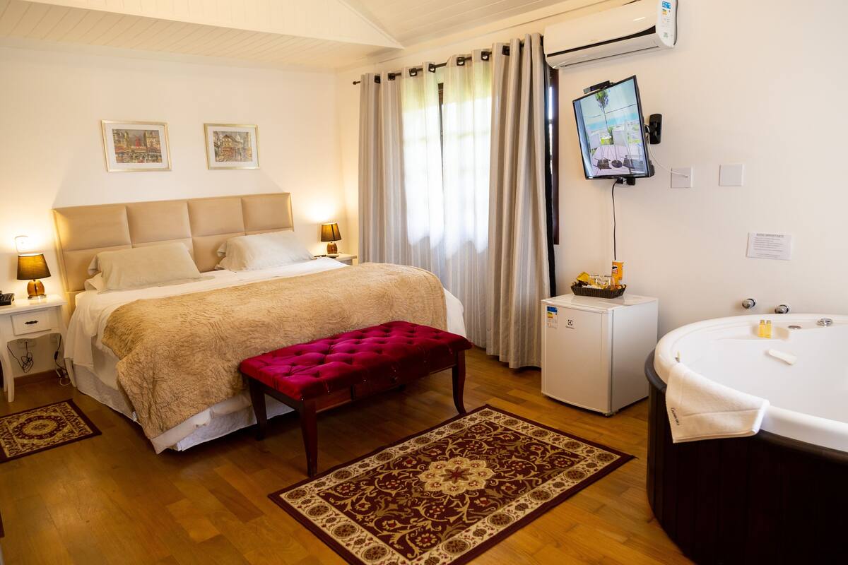 A hotel room with a large bed, a red bench, a small table with a lamp, a TV, a mini fridge, and a bathtub.