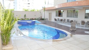 A swimming pool with lounge chairs and a hot tub, surrounded by plants and a building with large windows.