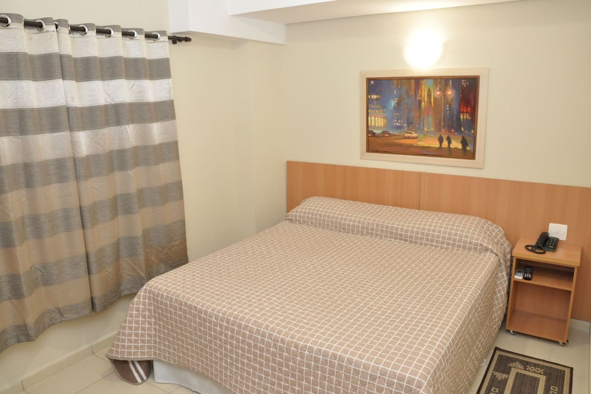 Una habitación de hotel con una cama, una mesita de noche, un cuadro en la pared y una cortina a rayas.