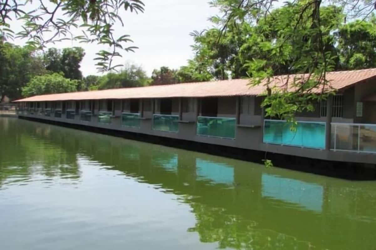 A long, narrow building with a red roof, situated by a calm body of water, surrounded by greenery.