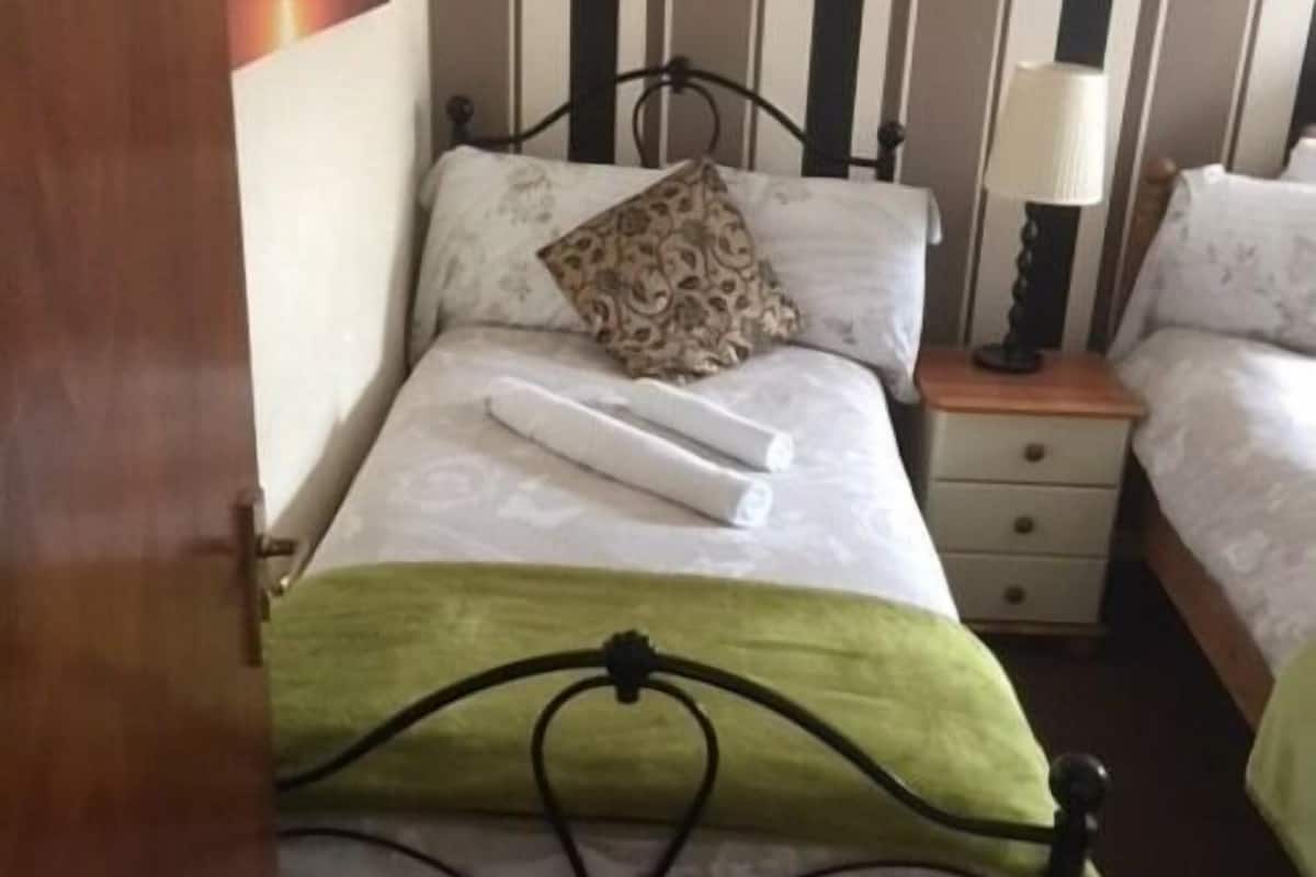 Una cama individual con una manta verde, un cojín estampado y una toalla blanca enrollada. Se observa una mesita de noche con una lámpara y un cajón.