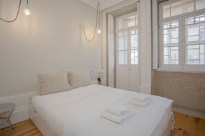 A neatly made bed with white linens and towels, a small table with a chair, and a hanging light fixture. - Liiiving in Porto - Bolhão Market Apartment II (Porto)