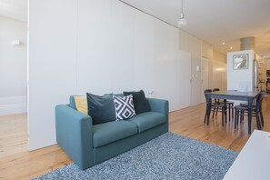 A modern living room with a teal sofa, patterned rug, and a dining area with a table and chairs. - Liiiving in Porto - Bolhão Market Apartment II (Porto)