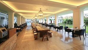 A spacious indoor dining area with long wooden tables and chairs, a grill station, and a view of greenery outside.