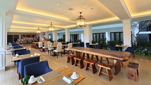 A spacious dining area with long wooden tables and blue upholstered chairs, featuring a modern chandelier and a view of the outdoors.