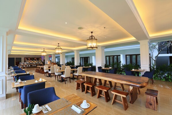 A spacious dining area with long wooden tables and blue upholstered chairs, featuring a modern chandelier and a view of the outdoors. - Grand Palace Hotel Sanur - Bali (Denpasar)
