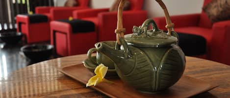 Een groene keramieken theepot met een bamboehandvat op een houten dienblad, geplaatst op een tafel in een kamer met rode stoelen en een zwarte kom op de achtergrond.
