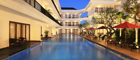 Una piscina con sillas de descanso y sombrillas, rodeada por un edificio de varios pisos con balcones y ventanas amplias.