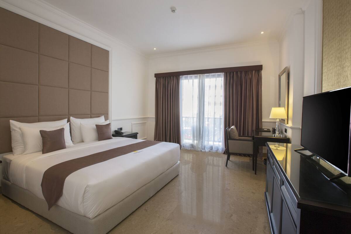 Une chambre d’hôtel avec un grand lit, un bureau, une chaise, une télévision et une fenêtre avec des rideaux.