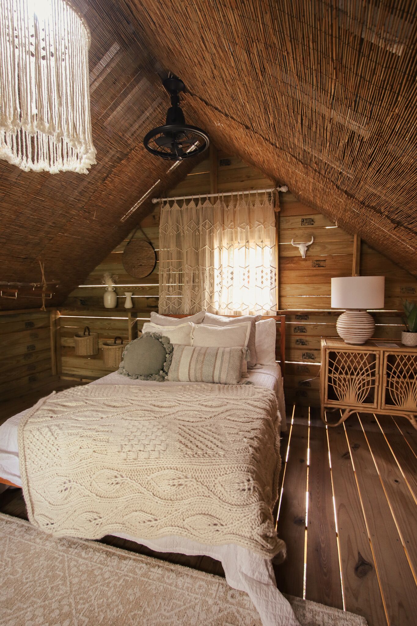 An attic bedroom with a sloped ceiling, a bed with a knitted blanket, a hanging macrame light fixture, and a rustic wooden wall with a cow skull decoration.