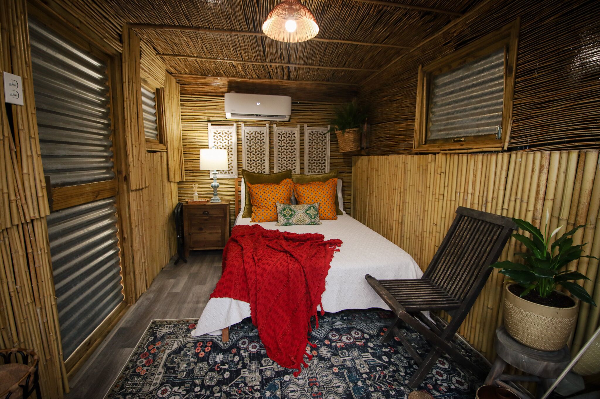 A compact room with a bed, a chair, a nightstand, a lamp, and a potted plant.