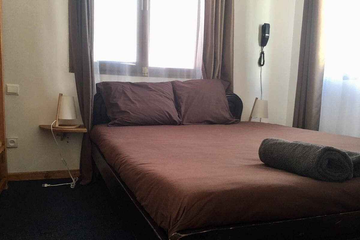 A hotel room with a bed, a bedside table, a lamp, a phone, and a window with curtains.