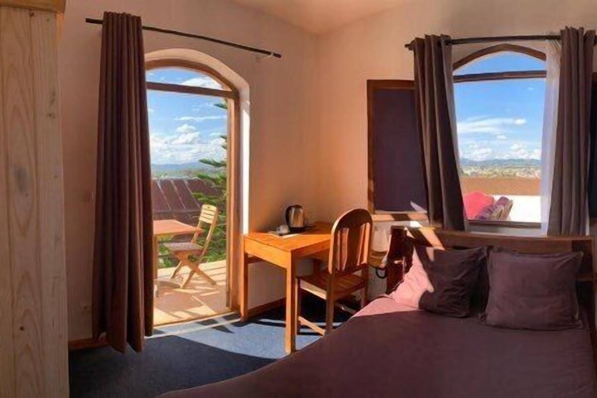 A bedroom with a bed, a desk, and a chair. There are two windows with curtains, one of which has a view of the outdoors.