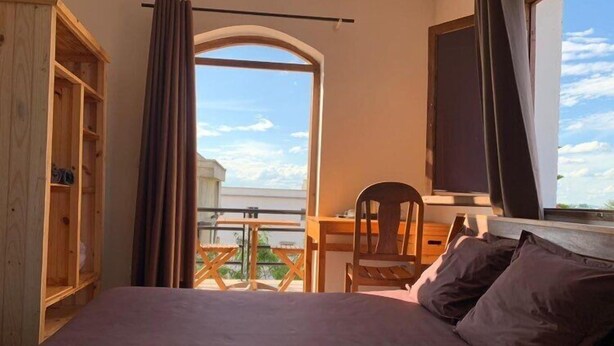 A bedroom with a bed, a desk, and a chair. There is a balcony with a view of the sky and clouds. - Iarimbato Hotel (Antananarivo)