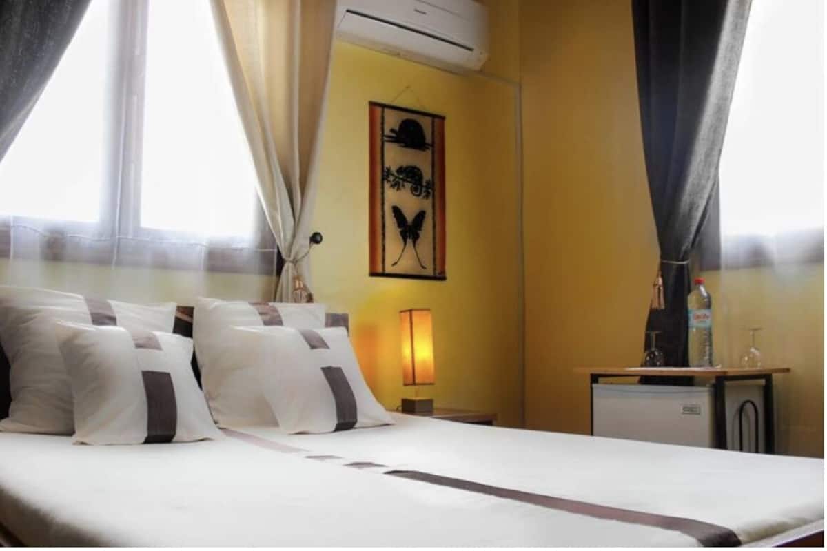 A hotel room with a bed, curtains, a wall-mounted air conditioner, a small table with a bottle and glasses, and a wall art piece.