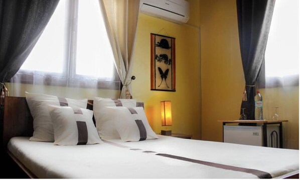 Habitación de hotel con cama, cortinas, aire acondicionado instalado en la pared, una mesita con una botella y vasos, y una obra de arte en la pared.