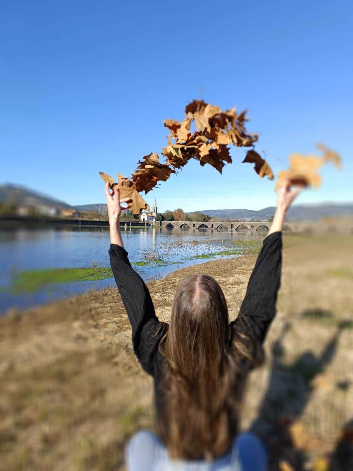 Une personne tenant des feuilles d’automne devant un ciel dégagé, un pont et une étendue d’eau.