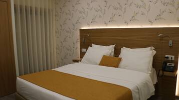 Habitación de hotel con una cama grande, cabecera de madera, mesita de noche con teléfono y ventana con persianas verticales.