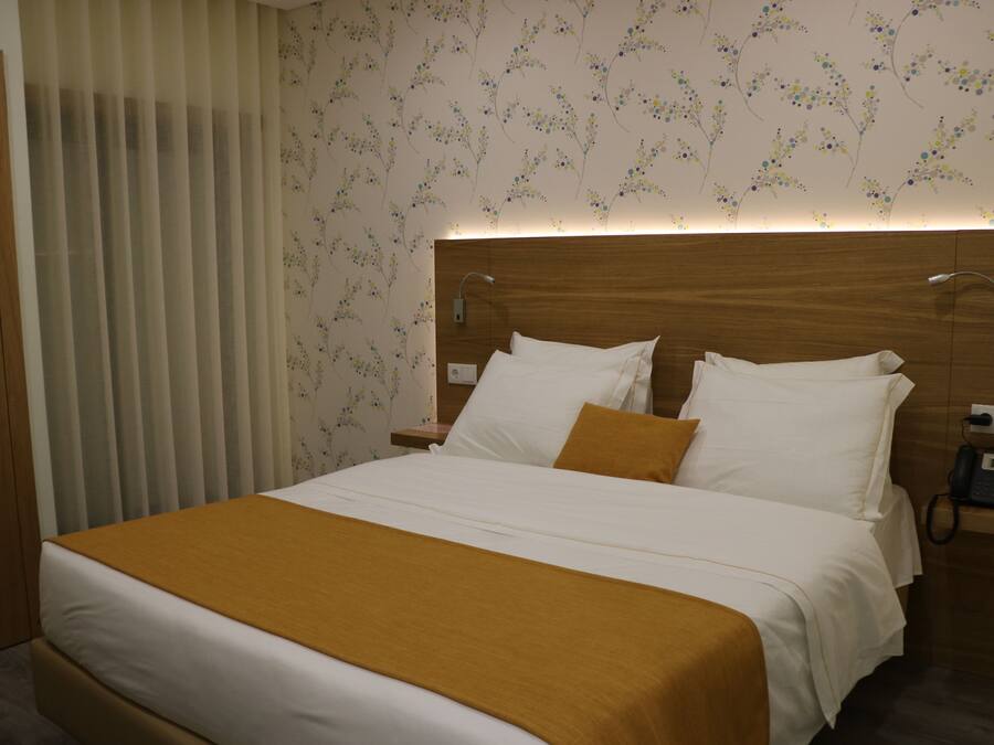Habitación de hotel con una cama grande, cabecera de madera, mesita de noche con teléfono y ventana con persianas verticales.