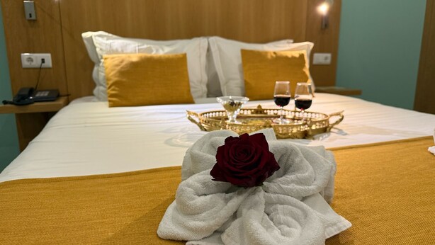 Una cama bien hecha con un edredón dorado, una toalla blanca con una rosa roja y dos copas de vino tinto sobre una bandeja.