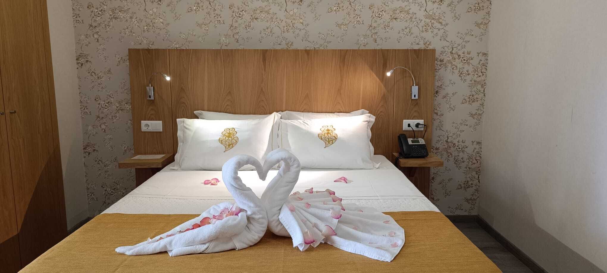 Habitación de hotel con cabecera de madera, papel tapiz floral, cama con ropa de cama blanca, dos almohadas con diseños de corazones, una presentación de toallas con forma de cisne y teléfono en la mesita de noche.