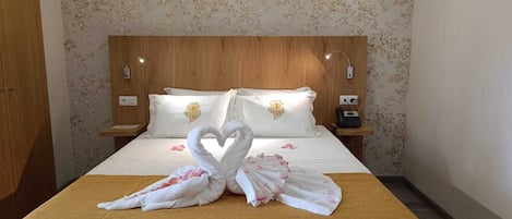 Habitación de hotel con cabecera de madera, papel tapiz floral, cama con ropa de cama blanca, dos almohadas con diseños de corazones, una presentación de toallas con forma de cisne y teléfono en la mesita de noche.