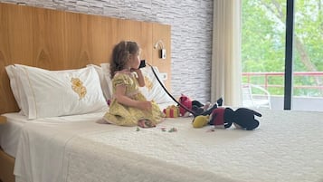 Un enfant est assis sur un lit dans une chambre d’hôtel. On peut voir une tête de lit en bois, une fenêtre donnant sur des arbres et une valise posée sur le sol.