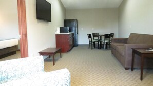 A living room with a sofa, a small table, a TV mounted on the wall, a refrigerator, and a dining area with chairs. - Edgewater Hotel (Put-in-Bay)