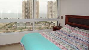 A bedroom with a large window, a bed with patterned bedding, a nightstand, and a hanging lamp. - Departamento Marina Horizonte 1 (Coquimbo)