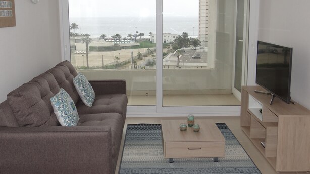 A living room with a brown sofa, a coffee table with decorative jars, a TV on a wooden stand, and a large sliding glass door with a view of the beach and palm trees. - Departamento Marina Horizonte 1 (Coquimbo)
