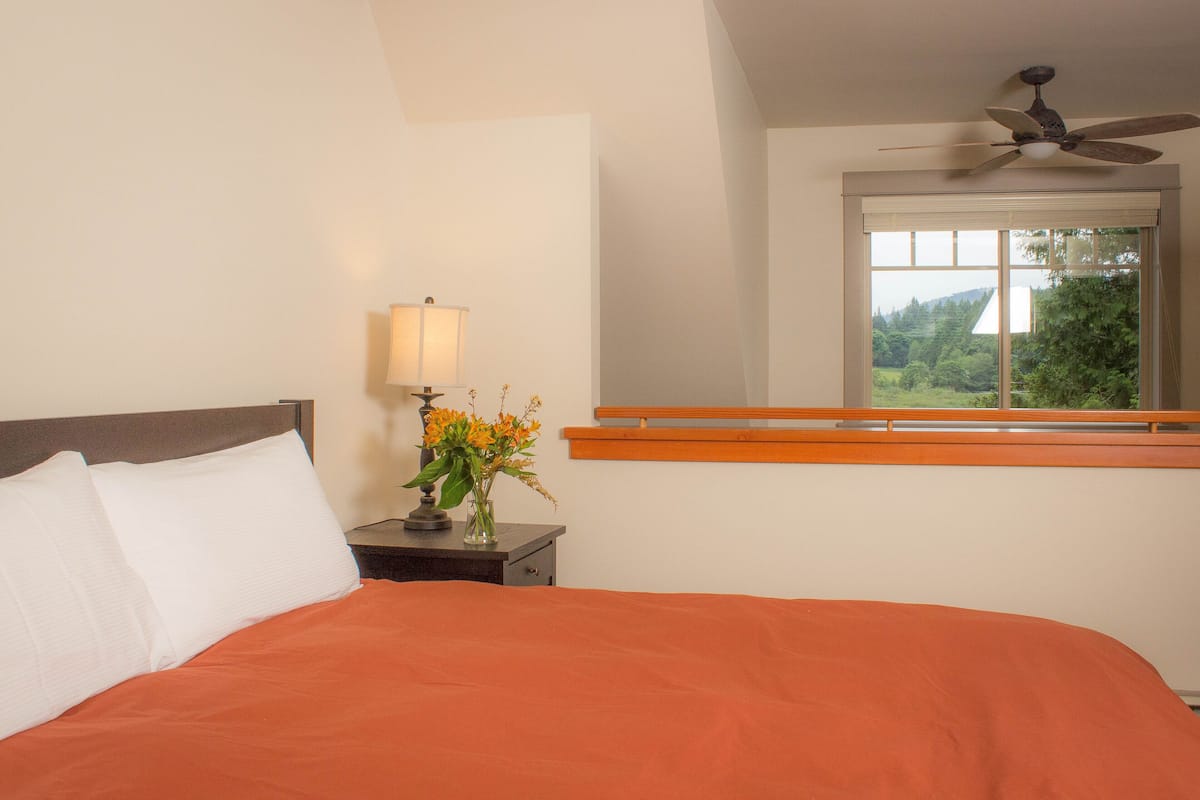 A hotel room with a bed, a nightstand with a lamp and flowers, a ceiling fan, and a window with a view of greenery.
