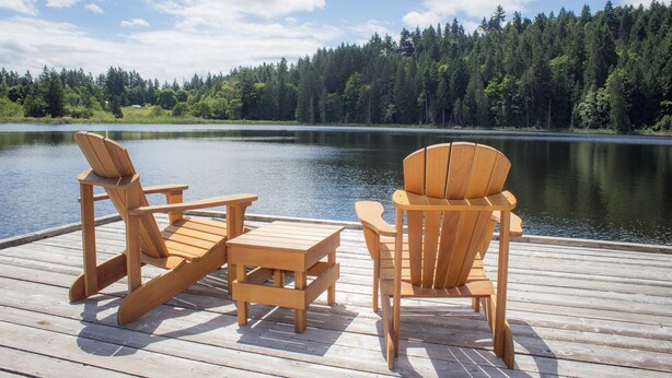 A wooden deck with two Adirondack chairs and a small table, overlooking a calm lake and dense forest. - The Cottages on Salt Spring Island (Salt Spring Island)