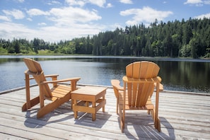 A wooden deck with two Adirondack chairs and a small table, overlooking a calm lake and dense forest. - The Cottages on Salt Spring Island (Salt Spring Island)