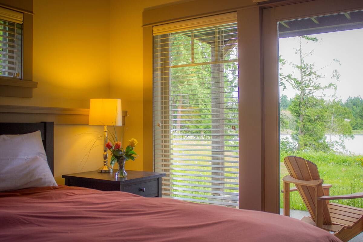 A bedroom with a bed, bedside table, lamp, and a view of a wooden chair outside.
